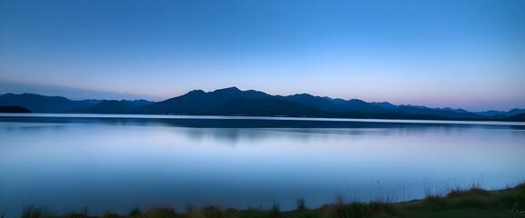 Fototapeta premium A landscape shot at dusk by the edge of a lake. Mountains on the horizon and the slightly blurred sky reflect on the calm surface of the water.