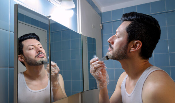 Hombre coloreando su barba con tintura de pelo frente 
a su reflejo en el espejo de ba&ntilde;o. Vista de perfil. 