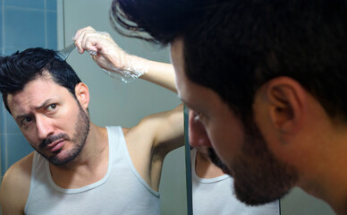 Hombre tiñendo su cabello y mirando su reflejo en el espejo de baño, usando guante de plástico y...