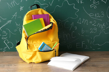 Backpack with different school stationery on wooden table against green chalkboard