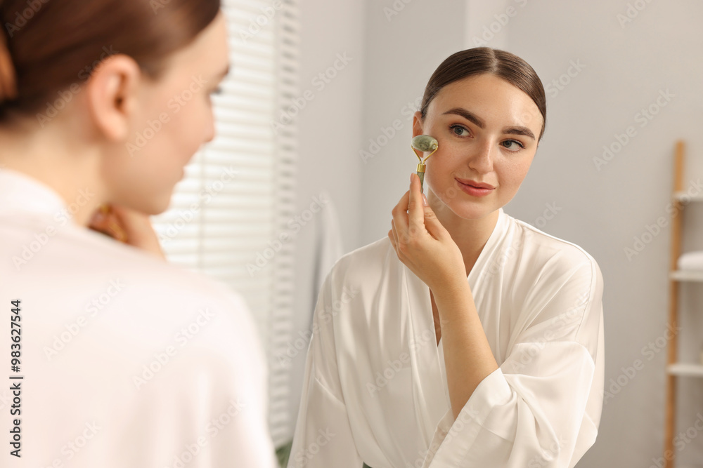 Sticker Beautiful young woman doing facial massage with roller near mirror at home