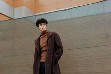 Fashionable young man in brown coat and turtleneck sweater standing with hands in pockets in front...
