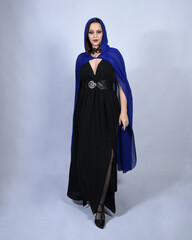 Full length portrait of  female red head model wearing  glamorous black witch dress with blue fantasy cloak halloween costume.  standing pose with gestural arms, isolated figure studio background.