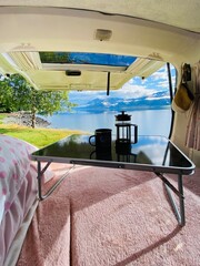 Coffee cup and coffee plunger on the table in the campervan with beautiful views of lake and blue sky at the background 