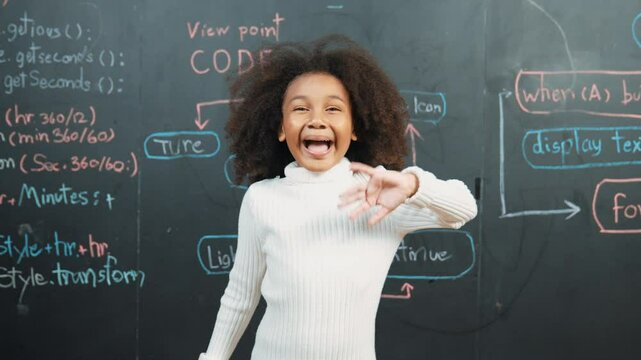 African girl smiling and waving at camera while standing at board with engineering prompt or coding, programing system written in STEM classroom. Child looking at camera while greeting. Erudition.