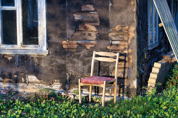 Abandoned weathered house exterior with rustic wooden chair and overgrown greenery in sunlight