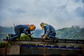 Two construction workers welding steel beams on a building structure, with a mountainous landscape in the background. Concept of teamwork, hard labor, and construction in challenging environments