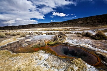 A geothermal hot spring surrounded by dry land and distant hills under a cloudy sky.