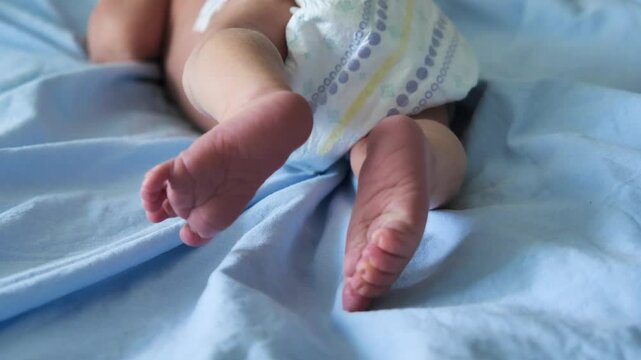 A baby lying on its stomach. Close-up of legs, creased feet and small toes. The baby is wearing a patterned nappy and lying on a blue sheet.