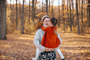 The mother, wearing a light grey sweater is holding daughter dressed in a orange sweater, smiles warmly as they share an affectionate embrace surrounded by colorful fallen leaves and tall trees. 