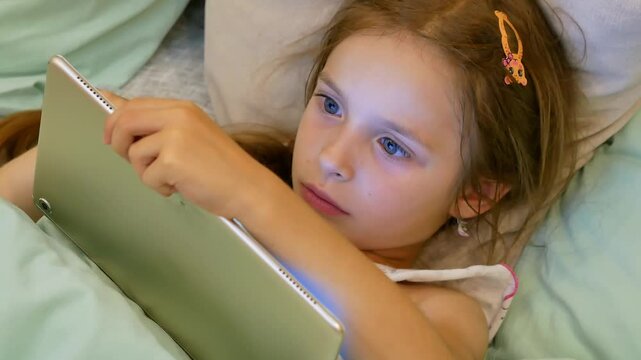 A little girl is lying in a crib holding a tablet in her hands, playing on a tablet or watching a video carefully. Blue eyes. 
