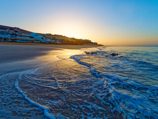 A beautiful sunrise over a sandy beach in Morro Jable on island Fourteventura. The sun is rising over a calm ocean, and the sky is a clear blue. There are palm trees and buildings in the distance.