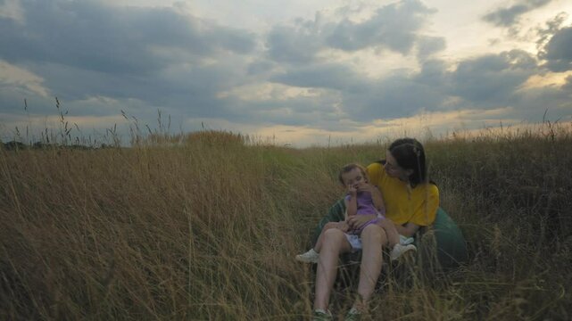 Loving charming mother sitting on beanbag chair, holding on laps cute toddler baby daughter,, bonding and relaxing in summer nature ,expressing happiness and positivity during vacations at countryside