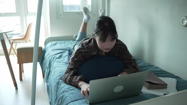 Asian girl prone on bed and typing on laptop