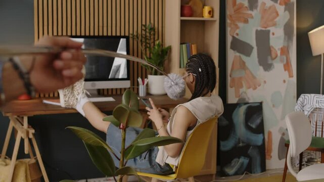 Biracial gen z girl sitting with her legs on desk in modern apartment while playful boyfriend teasing her with pp duster