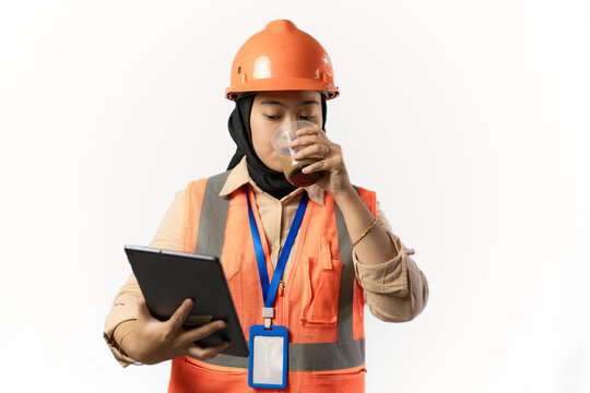 Young Indonesian female construction worker in hijab enjoying a cup of coffee while carrying a tablet, industrial and construction concept, isolated white background.