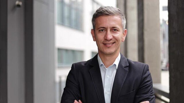 A confident and successful businessman in his 40s or 50s smiles at the camera with his arms crossed. He is standing in a modern office building, conveying experience, leadership, and success.