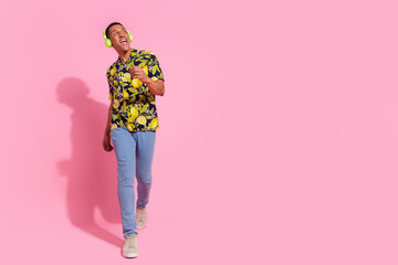 Full body photo of young man look empty space walk listen music headphones dressed tropical print clothes isolated on pink color background