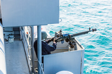 machine gun on a battleship. A marine holds a weapon. securing a ship. protection from pirates. a sailor in a black uniform is on automatic weapons duty on a battleship.