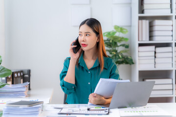 Focused and Determined: A young businesswoman in a green shirt, diligently working at her desk, multitasks, holding documents and speaking on the phone. She is surrounded by paperwork and a laptop, em