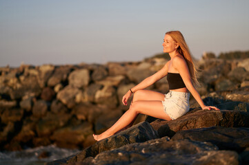 Beautiful young woman sitting on a rock near the sea. A lonely girl sits on the rocks by the sea and looks into the distance. Smiling blonde-haired woman sits on rocks by the sunset sea.