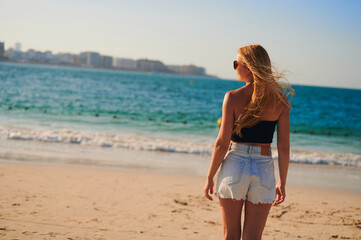 Fototapeta premium Young blonde European woman watches the sea waves from the back. A girl with a beautiful figure in shorts and top standing by the sea close-up.