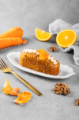 An appetizing carrot-orange cupcake decorated with cream and carrot chips lies on a plate, next to it there are carrots, oranges and nuts on a gray background. Baking concept for a coffee shop.