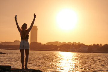 A golden sunset on the sea. A young woman with a beautiful figure standing by the sea with her hands up showing the V sign.