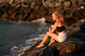 Woman sitting on rock and looking at bay. Beautiful European woman relaxing on the sea on summer holiday sitting on rocks.