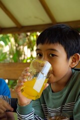 A young boy drinks fresh orange juice from a glass in an outdoor setting. The vibrant orange color of the juice complements the natural background, creating a refreshing, healthy moment of hydration.