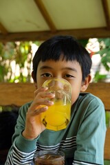 A young boy drinks fresh orange juice from a glass in an outdoor setting. The vibrant orange color of the juice complements the natural background, creating a refreshing, healthy moment of hydration.