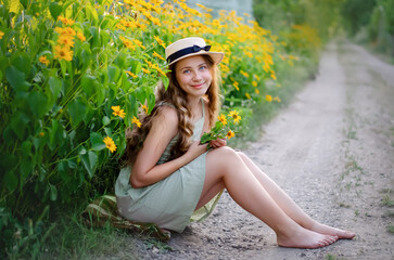 beautiful girl in a hat and dress smiles, sits on the ground in flowers