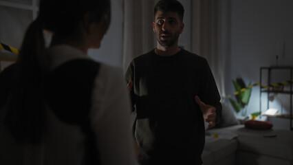 In a dimly lit indoor crime scene, a man gestures while talking to a woman, with yellow police tape...