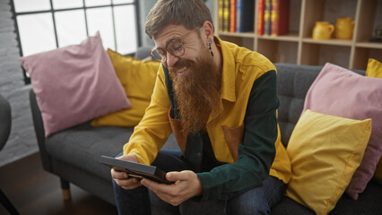 Smiling bearded man using tablet on cozy gray sofa with colorful pillows indoors