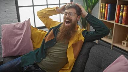 Fototapeta premium A cheerful bearded redhead man lounging on a couch indoors, expressing joy and comfort surrounded by books and plants.