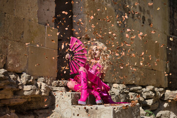 Drag queen, blonde, attractive, with crown, in pink dress and heeled boots, fanning herself with a big fan under a rain of dry leaves, sitting on stone steps. Concept pride, identity, diversity.