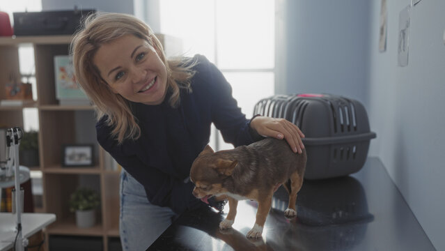 A young blonde woman in a veterinary clinic smiles while petting a chihuahua on a table, with a pet carrier in the background.