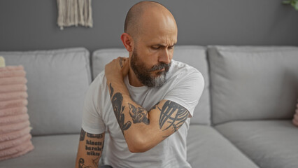 Fototapeta premium Handsome hispanic man with a beard and tattoos sits in a living room wearing a white shirt while holding his shoulder in pain
