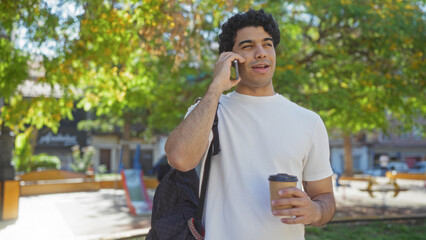 Handsome young hispanic man in an urban park enjoying a coffee while talking on his phone outdoors