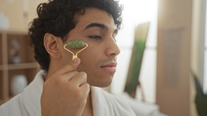 A handsome young hispanic man is using a jade roller on his face in a spa salon room for wellness.