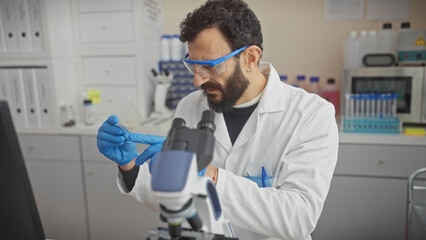 A bearded scientist examines a sample through a microscope in a laboratory setting, wearing blue gloves and protective eyewear.