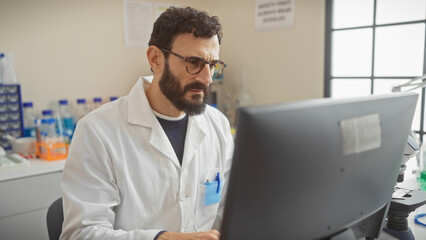 A mature man with a beard wearing glasses and a lab coat analyzes data on a computer in a research laboratory.