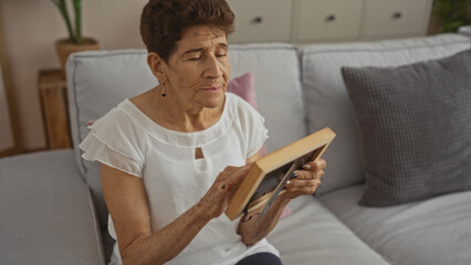 An elderly hispanic woman with short hair sits in her living room holding a picture frame, evoking...