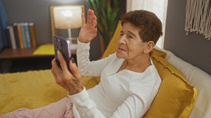 Elderly hispanic woman with short hair laying in bed at home, using a smartphone for a video call in a cozy bedroom.