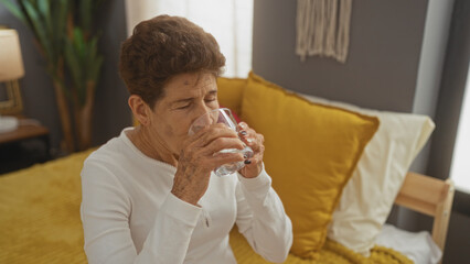 An elderly hispanic woman with short hair drinks water while sitting on a bed in a cozy bedroom, showcasing a calm interior setting.