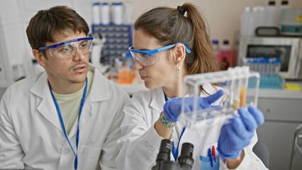 Two scientists, a man and a woman, examine samples in a laboratory setting, equipped with safety goggles and lab coats.