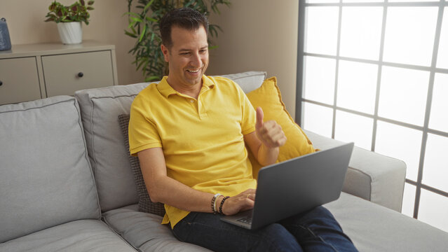 Hispanic man in a yellow shirt sitting on a couch in a living room, giving a thumbs up while using a laptop