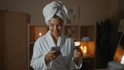 A young hispanic woman with a towel on her head, wearing a bathrobe, smiles while using her phone...
