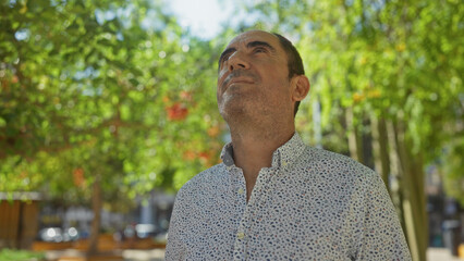 Mature hispanic man standing outdoors in an urban park, looking upwards with a thoughtful expression, surrounded by green trees and blurred city background.