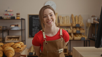 Young woman with red hair smiles warmly while working in a cozy bakery shop filled with fresh bread and pastries.
